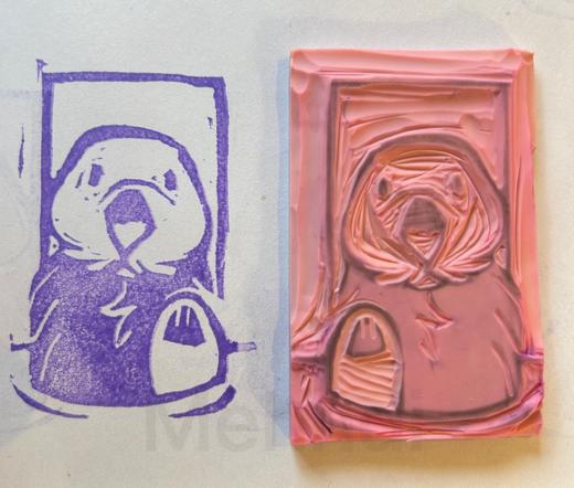 A picture of a purple stamp of a sea otter. They have their left paw up out of the water. Beside it is a pink block used to make the purple otter stamp, it is the same otter in a mirrored pose.