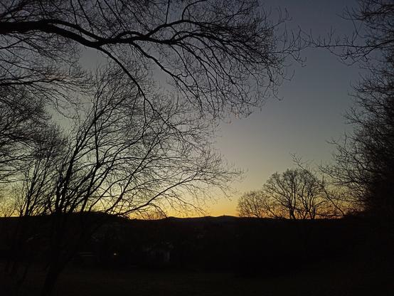 Blick Richtung Alarmstange und Köppel nach Sonnenuntergang. Unteres Bildviertel schwarz, darüber Höhenzug mit den beiden genannten Türmen mit gelblich Licht dahinter, selbiges über gehend nach oben in bläulichen wolkenfreien Himmel. Im Vordergrund quasi das Bild rahmend Sträucher und Astwerk.