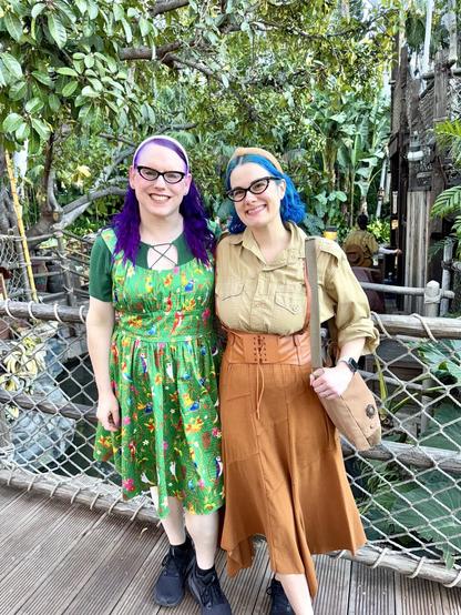 The image features two women standing side-by-side, posing for the camera against a lush green backdrop. The woman on the left has vibrant purple hair styled in pigtails, complemented by a green dress adorned with colorful flowers and leaves. Her counterpart on the right sports blue hair tied back in a ponytail, paired with a tan blouse and brown skirt.

In the background, a wooden bridge or walkway is visible, flanked by trees and foliage that creates a natural ambiance. The overall setting appears to be an outdoor location, possibly a park or garden, where the two women are enjoying each other's company amidst the serene surroundings.