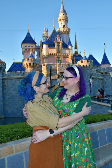 The image depicts two women embracing in front of the iconic Disneyland castle, their arms wrapped around each other's waists as they face one another.
The woman on the left has striking blue hair and is dressed in a tan shirt paired with an brown skirt. Her counterpart on the right boasts vibrant purple locks and wears a green dress adorned with colorful flowers. Both women wear headbands, adding a touch of elegance to their outfits.