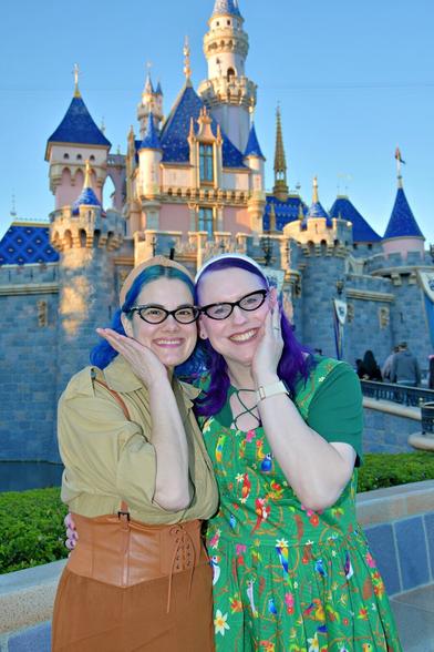 The image features two women posing for a photograph in front of the iconic Disneyland castle. The woman on the left has striking blue hair and is dressed in a beige shirt, brown skirt, and brown belt. Her companion on the right boasts vibrant purple locks and wears a green dress adorned with colorful flowers.

In the background, the majestic Disneyland castle rises against a brilliant blue sky, its towers reaching towards the heavens. The overall atmosphere exudes joy and excitement, capturing the essence of a magical day at the Happiest Place on Earth.