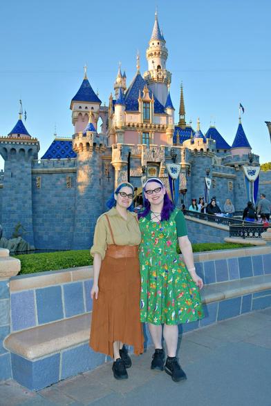 The image depicts two women standing side-by-side in front of the iconic Sleeping Beauty Castle at Disneyland, captured from an angle that showcases its majestic facade.

Both women are dressed in vibrant attire: one wears a green dress adorned with red and yellow flowers, while the other sports a brown skirt paired with a light-colored blouse. The woman on the left has her hair styled in a bun, whereas the woman on the right sports purple locks cascading down her back. They both don black shoes.

In the background, the castle's grandeur is accentuated by its blue roof and stone walls, set against a clear sky that adds to the enchanting atmosphere of the scene.