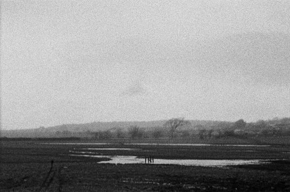 A black and white photo of a field. A small body of water sits in the middle of it