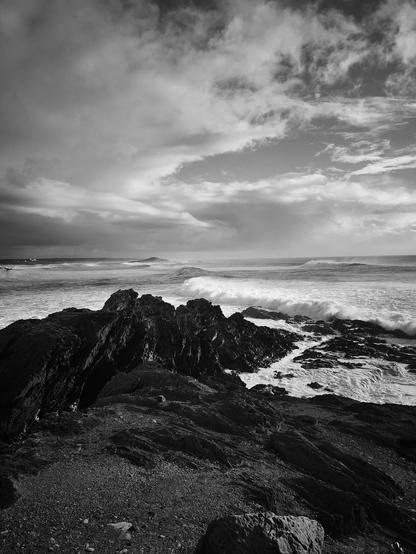 This black and white image features a dramatic coastal scene. In the foreground, there are rugged rocks and a rocky shoreline. Waves crash against the rocks, creating a dynamic contrast with the dark rocks and the white foam. The sky is filled with clouds, adding to the dramatic and moody atmosphere of the scene.