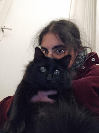 Moi avec les cheveux ébouriffés (je me suis levé à 6h du mat pour 5h de train, on m'excusera 😂) en train de tenir d'une main mon bon gros chat noir, Changiz, face caméra, avec ses grands yeux ébahis.