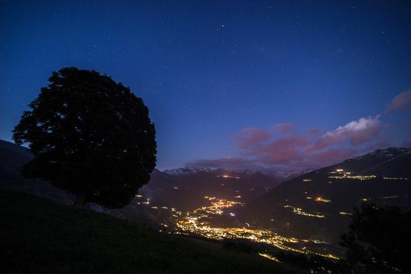 Photo d'un paysage de montagne nocturne.
Au premier plan à gauche, un arbre au tronc court et à la silhouette ronde semble regarder la vallée en contrebas. 
Le fond de la vallée est violemment éclairé par les lumières des villes, des villages illuminent aussi les flancs des montagnes. Quelques nuages, rendus roses par la pollution lumineuse, s'accrochent aux sommets saupoudrés de neige.
Le ciel est bleu et parsemé d'étoiles.
-
Photo of a mountain landscape at night.
In the left foreground, a tree with a short trunk and round silhouette seems to look down on the valley below. 
The valley floor is brightly lit by city lights, while villages light up the mountainsides. A few clouds, made pink by light pollution, cling to the snow-dusted peaks.
The sky is blue and dotted with stars.