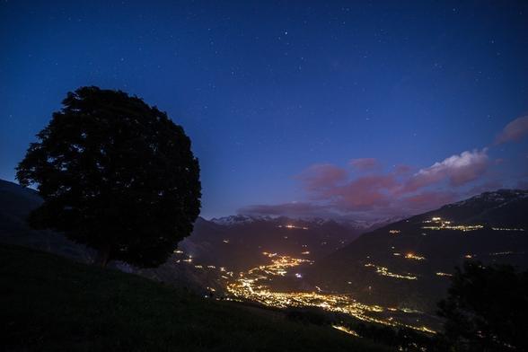 Photo d'un paysage de montagne nocturne.
Au premier plan à gauche, un arbre au tronc court et à la silhouette ronde semble regarder la vallée en contrebas. 
Le fond de la vallée est violemment éclairé par les lumières des villes, des villages illuminent aussi les flancs des montagnes. Quelques nuages, rendus roses par la pollution lumineuse, s'accrochent aux sommets saupoudrés de neige.
Le ciel est bleu et parsemé d'étoiles.
-
Photo of a mountain landscape at night.
In the left foreground, a tree with a short trunk and round silhouette seems to look down on the valley below. 
The valley floor is brightly lit by city lights, while villages light up the mountainsides. A few clouds, made pink by light pollution, cling to the snow-dusted peaks.
The sky is blue and dotted with stars.