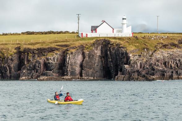 Two people kayaking in a yellow tandem kayak on calm waters near rocky cliffs, with a white lighthouse and cottage in the background atop a grassy hill in Dingle Harbour, County Kerry, Ireland.