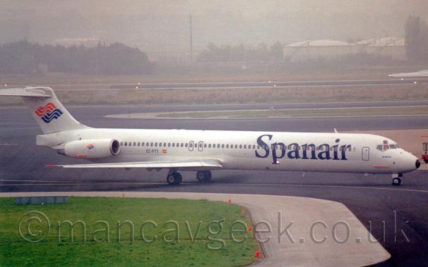 Side view of a white, twin engined jet airliner with the engines mounted on the sides of the rear fuselage, taxiing from left to right along a black taxiway.
The plane has large, blue, billboard-style "SpanAir" titles on the forward fuselage, and orange and blue waves on the tail, a design repeated on the engine pods.
The registration "EC-FTT" is on the lower rear fuselage in black.
Large patches of grass with grey tarmac surrounds line the sides of various taxiways in the foreground and background, with trees and large grey fuel tanks in the distance, under murky grey skies.