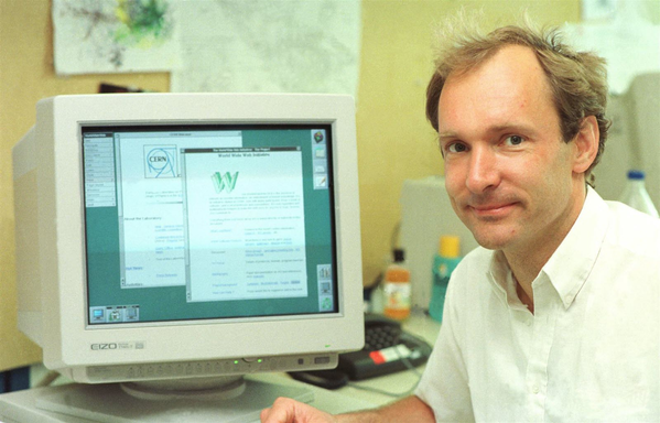Tim Berners-Lee looking to the camera with a desktop computer behind him. The computer has open web pages on the desktop