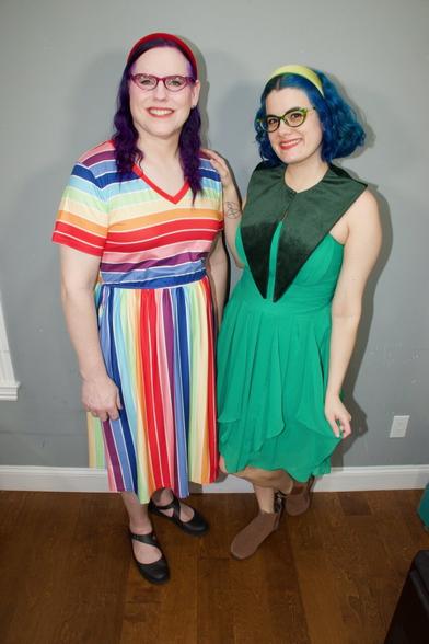 The image features two women standing side-by-side against a gray wall, posing for the camera.

On the left, a woman with dark purple hair styled in a headband stands out in a vibrant rainbow-striped dress. Her attire is complemented by black Mary Jane shoes and a subtle smile as she gazes directly at the camera.

To her right, another woman sports short blue hair and a green sleeveless dress with a deep V-neckline. She wears brown flats and also faces the camera with a gentle smile.

The background of the image consists of a gray wall, providing a neutral backdrop for the two women to take center stage.