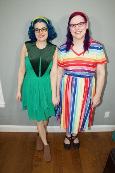 The image features two women standing side-by-side against a grey wall, facing the camera. The woman on the left has dark blue hair styled in a bob with bangs, complemented by a yellow headband and black-framed glasses. She wears a green dress with a V-shaped neckline and brown ankle boots.

In contrast, the woman on the right sports purple hair, also styled in a bob with bangs, paired with red-framed glasses. Her attire consists of a vibrant striped shirt featuring a mix of red, orange, yellow, blue, and white hues, along with black Mary Jane shoes.

The background of the image is a grey wall with white trim at the bottom, while the floor appears to be made of hardwood or laminate material.