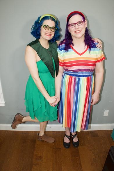 The image features two women standing side-by-side against a gray wall, their arms around each other's shoulders.

The woman on the left has short blue hair and is dressed in a green dress with brown shoes. The woman on the right has long purple hair and wears a vibrant striped dress with black heels. Both individuals are smiling at the camera, exuding warmth and camaraderie.

In the background, a gray wall provides a neutral backdrop for the subjects, while the wooden floor adds a touch of natural elegance to the scene. Overall, the image captures a heartwarming moment between two friends or family members, radiating positivity and joy.
