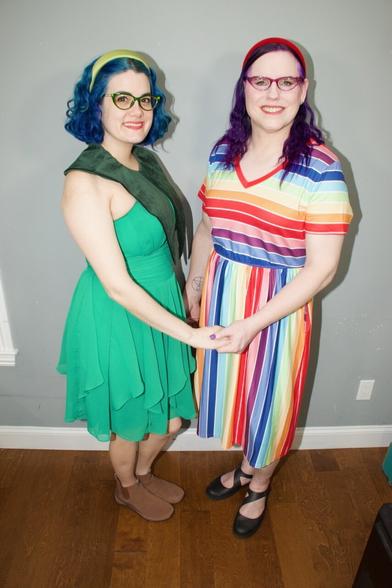 The image depicts two women standing side-by-side, facing the camera, with their hands clasped together in front of them.

The woman on the left has short blue hair and is wearing a green dress that falls just above her knees. She also wears brown shoes and has a dark green scarf draped over her shoulders. The woman on the right has long purple hair and is dressed in a vibrant, striped dress featuring red, orange, yellow, blue, and white hues. Her outfit is completed with black flats.

The background of the image features a gray wall with white trim at the bottom, while the floor appears to be made of hardwood or laminate material.