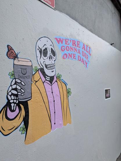 A mural of a skeleton dressed in a yellow suit holding up a beer that reads "Darkness" instead of "Guinness." The text on the mural reads, "We're all gonna die one day."