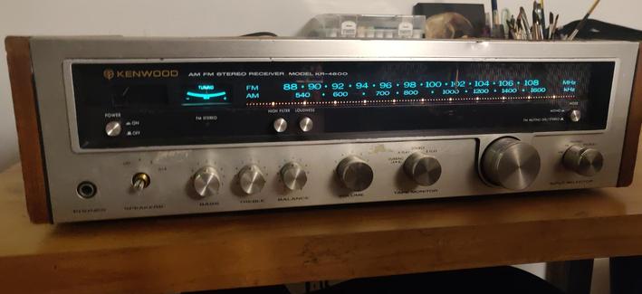 A mid-70s Kenwood AM/FM Stereo receiver. It has a silver front panel with several silver knobs, woodgrain panels on either side, and a wide black window spans the width of the unit for the turquoise-lit AM/FM tuner.