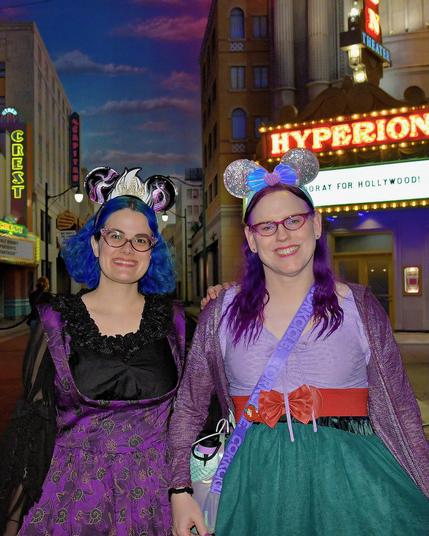 The image depicts two individuals standing side by side against an urban nighttime backdrop. Both are wearing costumes that include large, stylized ears and glasses, which are characteristic of characters from the "Harry Potter" series. The person on the left has blue hair styled in a bob with a black headband featuring white accents, while the individual on the right has purple hair styled in long dreadlocks with a pink headband. They are both wearing purple and green dresses that have floral patterns. Both individuals are smiling and appear to be enjoying themselves.

In the background, there is a building with illuminated signs, one of which reads "HYPERION" in large red letters. The sky above them is dark with hints of blue, suggesting it's either dusk or nighttime. There are no visible texts other than the signage and the costumes that provide context to their attire.

The image seems to be a playful representation of fans at a Harry Potter event or convention, possibly taking pl