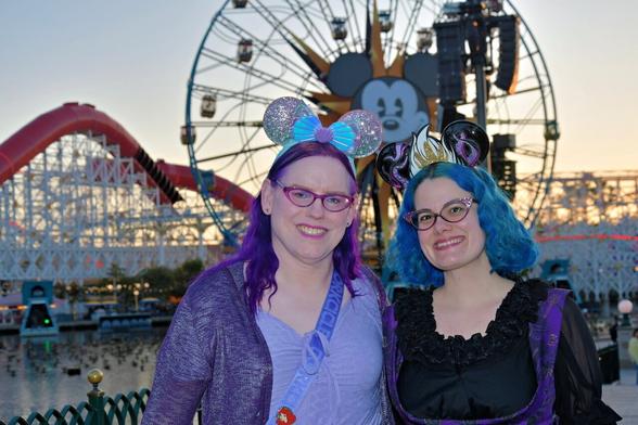 The image depicts two individuals standing together at what appears to be an amusement park during twilight hours. The person on the left is wearing a purple top, green skirt, and has blue hair styled in buns. They are holding a water bottle with a strap over their shoulder. The individual on the right is dressed in a black outfit with a purple trim, also sporting blue hair styled in buns. Both individuals are wearing glasses and appear to be smiling at the camera.

In the background, there's a large Ferris wheel with illuminated figures that resemble Mickey Mouse and Minnie Mouse, suggesting this could be a Disney theme park. The sky is transitioning from daylight to nighttime, with hues of orange and blue blending into each other. There are no visible texts or discernible brands in the image.

The photograph captures a moment of leisure and enjoyment, likely intended to convey the fun and excitement associated with visiting an amusement park. The lighting conditions suggest that thi