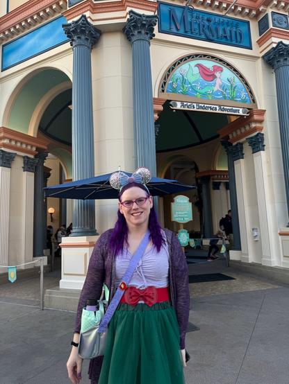 The image shows a person standing outside what appears to be an entrance to a themed attraction, possibly a theme park. The individual is wearing glasses, a purple headband with ears attached, and a green skirt. They are holding a blue umbrella in one hand and a silver bag in the other. The person's attire suggests they might be dressed for a costume or cosplay.

The building behind them has a classical architectural style with large columns supporting an arched entranceway. Above the entrance, there is a sign that reads "Mermaid," indicating the name of the attraction. The facade features decorative elements such as blue tiles and a mosaic-like design above the main entrance.

The ground appears to be paved, and there are no other people visible in the immediate vicinity of the person. The lighting suggests it is daytime with natural light illuminating the scene. There are no discernible texts providing additional context or information about the location or event.