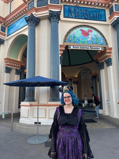 The image shows a person standing outside a building that appears to be part of a theme park or entertainment venue, given the signage "Mermaid" prominently displayed on the facade. The individual is wearing a black dress with purple sleeves and has blue hair styled in a bob cut. They are also wearing a crown-like headpiece that resembles a mermaid's tail fin, which complements their outfit. In front of them, there is an open umbrella providing shade. The building itself features classical architectural elements such as columns and arches, with a decorative mosaic or tile design above the entrance. There are people visible in the background, suggesting that this location is a public space where visitors can enjoy the attractions inside.