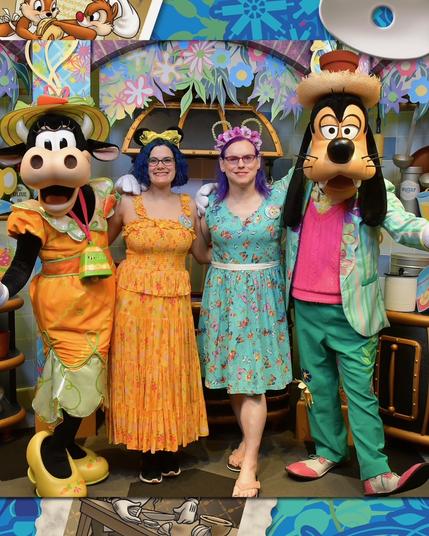 The image features two women posing alongside Disney characters Clarabelle and Goofy at a themed event.

Clarabelle is attired in an orange outfit, complete with a matching hat, while Goofy sports a light blue jacket over a pink shirt and green pants. The woman on the left wears a yellow sundress with a floral pattern, complemented by a purple flower crown. Her counterpart on the right dons a turquoise dress adorned with red flowers.

The background of the image is a vibrant mural featuring cartoon characters and plants, adding to the festive atmosphere of the event.