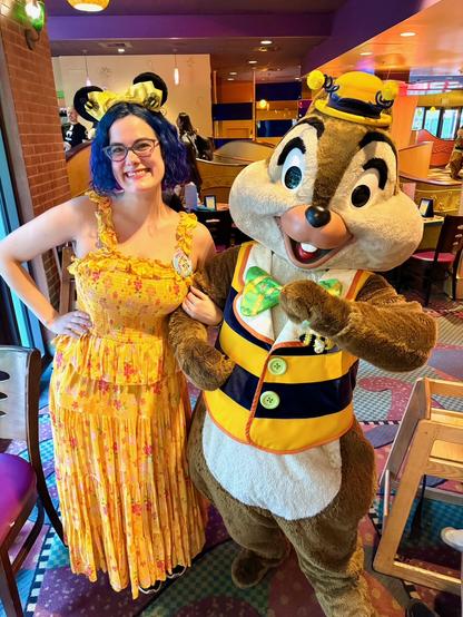 The image depicts a woman standing alongside a person dressed as Chip.

The woman has dark hair styled with bee themed mickey ears on top of her head, and she wears a yellow sundress with red flowers. She stands to the left of the character, facing forward with her right hand resting on the character's shoulder. The character is positioned slightly behind her, looking directly at the camera with their mouth open in a smile.

In the background, a restaurant or diner setting is visible, featuring tables and chairs.