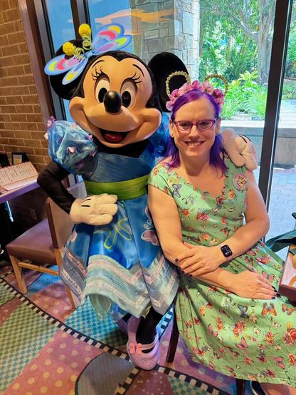The image features Minnie Mouse and a woman posing together for a photograph. 

Minnie is dressed in her signature blue dress adorned with white polka dots, complemented by a matching bow on her head. She has black ears and a large smile on her face.

The woman, positioned to the right of Minnie, wears a light green dress with pink flowers and has purple hair. Her arms are crossed over her lap as she sits in a chair.

In the background, a window offers a view of trees and bushes outside, while a brick wall is visible behind Minnie's left shoulder. The overall atmosphere suggests that this image was taken at an event or gathering where characters from Disney were present.