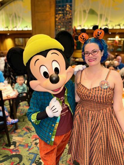 The image depicts a woman posing for a photograph alongside Mickey Mouse at Disney World. The woman has blue hair styled in an updo and is wearing a vibrant orange dress adorned with black stripes, complemented by a matching headband featuring two jack-o-lanterns on either side. She also wears a round pin on her left chest.

Mickey Mouse stands to the left of the woman, dressed in his signature yellow hat and blue shirt, paired with red pants. His right hand is raised in a thumbs-up gesture, while his left arm rests on the woman's shoulder.

In the background, several individuals are seated at tables, although their faces are not visible due to the distance from the camera. The atmosphere appears festive, with the presence of Mickey Mouse and the woman's attire suggesting that they may be celebrating Halloween or another holiday.