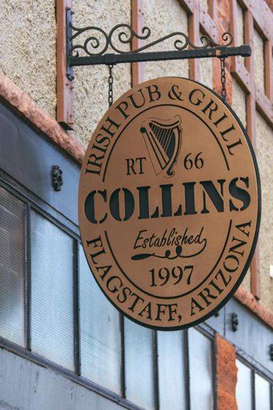 Hanging outdoor sign for an Irish restaurant in Flagstaff.  Over 25 years there now the sign is labeled.  Copper colored metal oval hangs from a black wrought iron bracket.  The letters and harp designs are cut through the top plate to expose the black one under it.  The rest of the building around this sign shows stucco, wood and red brick on the facade with windows set high at the storefront. Collins is the name.