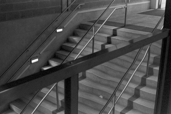 A view of a concrete, institutional staircase with brushed steel railings going downwards. The view is from above and through a metal banister, the view above is clear but, below the banister, the lines of the steps and railings are gently warped by the invisible glass panels.