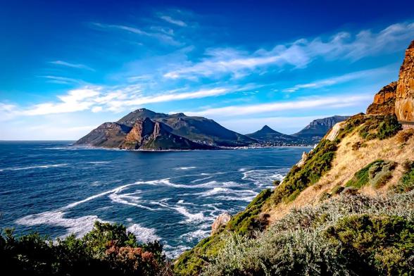 This image shows a coastal landscape with a large body of water, likely an ocean, meeting rugged cliffs and mountains in the distance. The foreground features green vegetation and rocky formations. The sky is bright with scattered clouds, creating a picturesque and serene natural scene.
