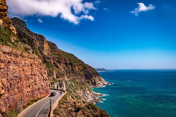 This image shows a scenic coastal road winding along steep cliffs with the ocean on one side. The cliffs are rugged and rocky, with some greenery. The sky is bright blue with a few clouds, creating a picturesque and dramatic landscape. There is a vehicle on the road, indicating that this is a route with vehicular access, offering stunning views of the coastline.
