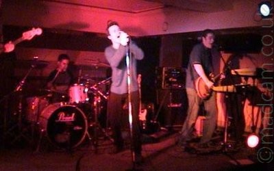 3 members of a band performing on a stage, on the left is the drummer at the back of the stage, with short dark hair and a grey and black t-shirt.
In the middle is a man with short dark hair, wearing a grey jumper and black trousers, leaning on a microphone stand as he sings into the mic.
On the right is a man with short dark hair, wearing  a grey t-shirt and dark trousers, playing an electric guitar, and singing into a microphone on a stand, behind a bank of keyboards.
The neck of a bass guitar can be seen right on the left of the frame, with a hand holding it half way along.
The room is lit by a group of red lights, with shadows reaching out in all directions.