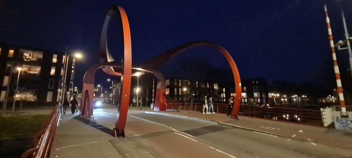 De Rode brug (red bridge) over the river Vecht at night. The draw-bridge consists of two red, metal u's. The one on the far side is flattened at the top, the other one resting on it, it's legs curving down in a big swoop towards the viewer and the one lane road, flanked by bike= and foothpaths on both sides, they are bolted to. In the background a line of four story apartment buildings with lit windows line de river. A car is entering the far side of the bridge, it's lights shining over it.