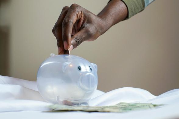 Une main droite introduit une pièce de monnaie dans une tirelire en forme de cochon. La tirelire est transparente.
