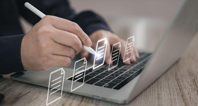 A stock image supposed to represent someone working on multiple documents; it's a close-up photo of a laptop with someone's hands positioned above the keyboard. The left hand is on the keys, the right is holding a stylus and tapping on some digital icons representing documents hovering above the keyboard.