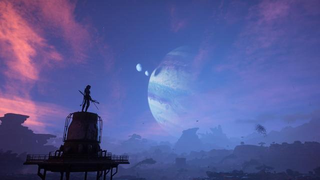 A video game photo capture, from Avatar: Frontiers of Pandora. A silhouetted Na’vi stands atop a dilapidated utility tower overlooking a sunset scene on Pandora, the gas giant Polyphemus with two additional moons in orbit, casting a shadow on the blue and purple clouds of the planet. Mountains and trees are seen on the horizon.