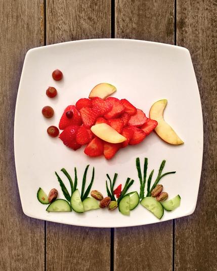 Ein Fisch gelegt aus Erdbeeren- und Apfelscheiben. Der Körper ist aus Erdbeeren, die Flossen aus Apfel gemacht. Luftblasen aus halben dunklen Weintrauben. Unten drunter Seegras aus Gurkenschale auf Gurkenscheiben und Pistazien.