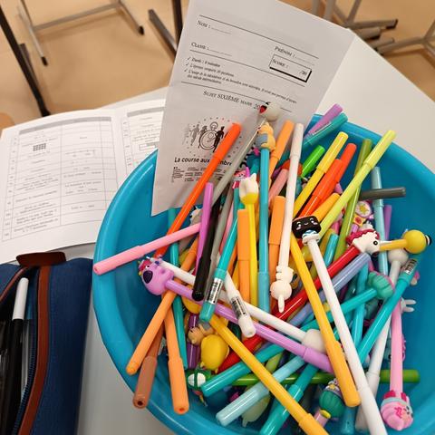 Un saladier rempli de stylos colorés avec des embouts fantaisie, posé sur une table avec des copies d’évaluation de mathématiques.