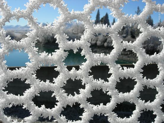 Closeup of thick hoar frost on a hexagonal wire fence (chicken wire), creating hexagons on hexagons on hexagons. Seen out-of-focus through the hexagonal pattern is a lake, with white frosted trees on its shoreline and snow-capped mountains behind. Lake Tekapo, New Zealand.