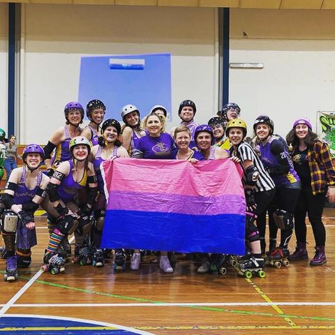 Lots of roller derby skaters in purple uniforms holding up a Bi Pride Flag. Emma is right in the middle behind the flag.