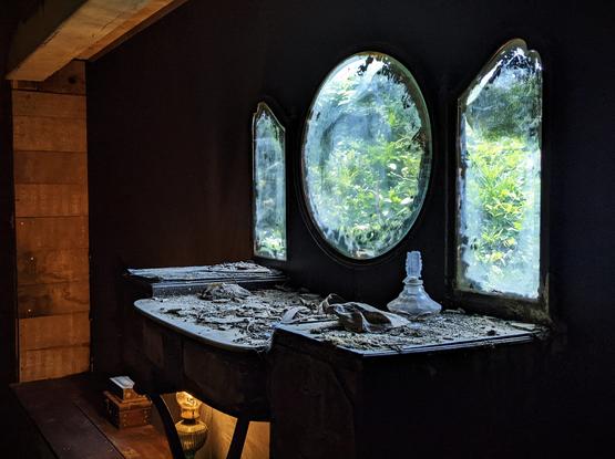 Part of an installation. An old window in three parts. Outside, some trees. Inside it's dark. In front of the window, an old desk with some objects, all covered in ash.