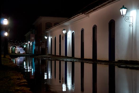 A cobblestone street of a historic center during night time. The middle of the street is filled with water, which reflects the light of the street lamps and the white colonial style buildings with colorful windows and doors.