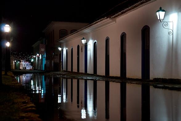 A cobblestone street of a historic center during night time. The middle of the street is filled with water, which reflects the light of the street lamps and the white colonial style buildings with colorful windows and doors.