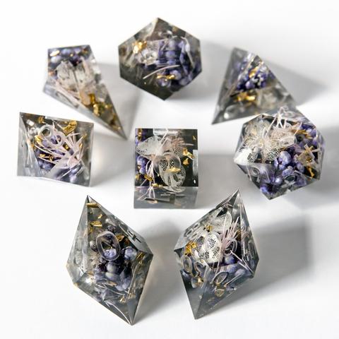 Ein Würfelset in Transparentem schwarz mit Blumen im Inneren. Das Set besteht aus einem D4-Shad, einer D4-Pyramid, D6, D8, D10, D100, D12 und D20. Das Set liegt auf weißem Untergrund mit der höchsten Seite nach oben. Die Würfel sind im Bild um den D6 angeordnet. Das Set ist noch Work in Progress und noch nicht geschliffen, poliert oder bemalt. Der D10 und der D00 sind im Vordergrund scharf dargestellt. Die anderen Würfel sind im Hintergrund leicht unscharf zu sehen.