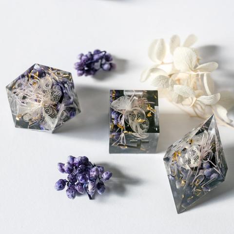 Ein D20, D6 und D00 auf weißem Untergrund. Außerdem sind auf dem Bild einige der Blumen zu sehen, welche in den Würfeln verarbeitet wurden.