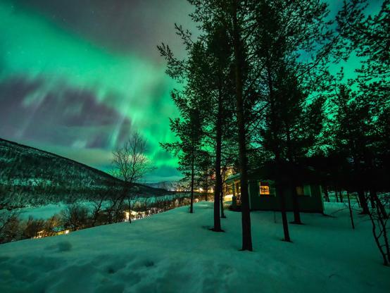 At night, under a starry sky lit in green by aurora. Ground is covered in snow, pine trees are dark silhouettes. On the right, amidst trees, a cabin has a window lit in yellow light, as is the village fare in the background at the center of the image.