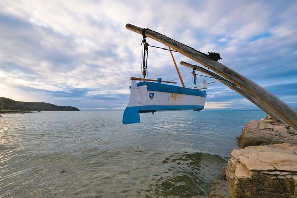 A blue and white boat is suspended on two masts. On the right you can see the stone jetty. The boat is suspended above the water. The sky is cloudy, but the sun is partially shining through.