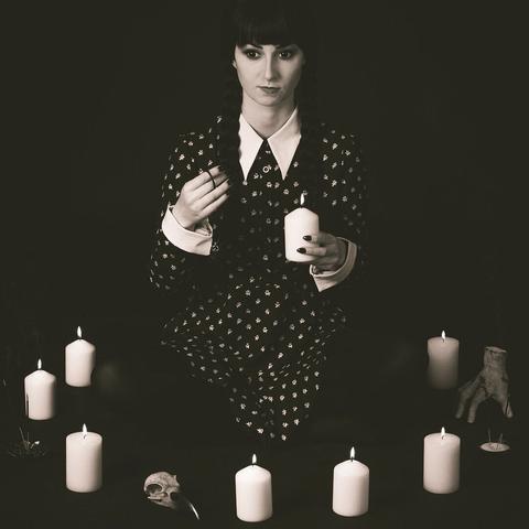 Cosplayerin als Wednesday Addams mit eiskaltem Händchen auf der Schulter sitzt im Kerzenkreis.