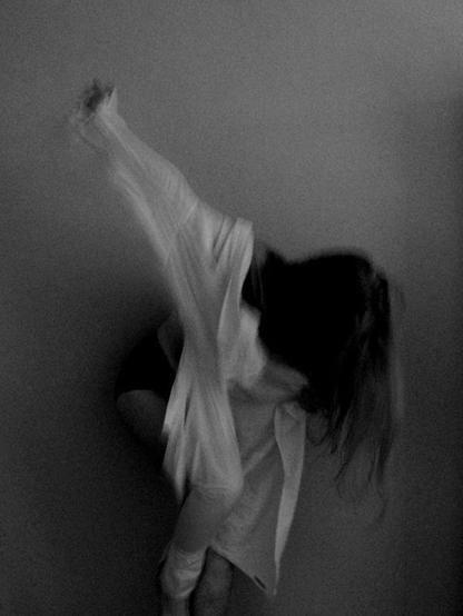 A woman is trying to put a shirt on but instead of putting her arms in the sleeves she is trying to get it on both backwards and with one arm in one sleeve and her right leg in the other one. Her right arm is holding the shirt up and she is leaning forwards since she can´t stand straight with the shirt put on like that. Her hair is haning down, hiding her face. The image is a bit blurry because of her motion during exposure.